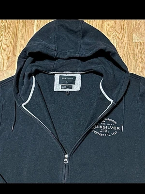 Svart zip-hoodie från Quiksilver - Svart hoodie med dragkedja från Quiksilver. Klassisk modell med huva, dragsnören och två fickor framtill. Tryckt logga på bröstet och mjukt material på insidan. Perfekt för en avslappnad streetstil.