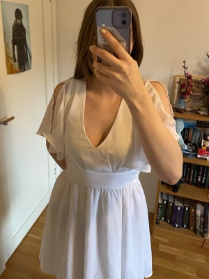 Studentklänning - Säljer en vit kortklänning med v-ringad front, axelbandslös design och luftiga, lösa ärmar. Klänningen har ett brett midjeband och knyt i ryggen för extra detalj. Utmärkt som studentklänning