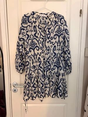 Vit och blå kortklänning med knyt - Kortklänning i vitt och blått med ett iögonfallande mönster. Klänningen har långa ärmar med resår vid ärmslut, v-ringning och knytband i midjan för en snygg passform. Tillverkad i ett lätt och luftigt material, perfekt för varmare dagar.