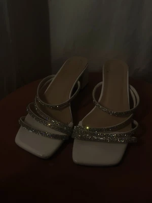 Vita sandaler med glittriga remmar - Säljer ett par vita sandaler med fyrkantig tå och flera tunna remmar täckta av glittrande stenar. Sandalerna har en låg, bred klack och öppen häl. Perfekta för dig som vill ha något extra till outfiten. Materialet är syntet med dekorativa detaljer.