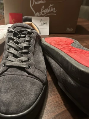 Gråa Christian Louboutin sneakers - Snygga gråa sneakers från Christian Louboutin i mocka med klassisk röd sula. Skorna har rund tå, gråa skosnören och låg profil. Perfekta för dig som vill ha något exklusivt och stilrent med en ikonisk touch.
