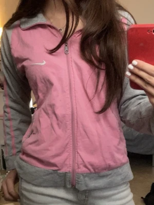 Rosa och grå jacka från Nike - Säljer en sportig rosa och grå jacka från Nike med dragkedja framtill. Jackan har långa ärmar och två fickor. Perfekt för en avslappnad stil eller träning. 🩷