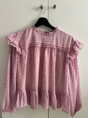 Rosa blommig blus från Lindex - Söt rosa blus från Lindex med små blommor i rosa och blå. Blusen har volangdetaljer vid axlar och nederkant, samt långärmad design med resår vid ärmslut. Tillverkad i ett lätt och luftigt material, perfekt för en trendig och romantisk look. Använd 1-2 gånger så fortfarande så gott som ny😊
