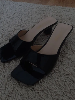 Svarta lackade klackade sandaler - Säljer ett par svarta sandaler med lackad finish och bred rem över foten. De har fyrkantig tå och klack, samt öppen häl. Snygg och trendig modell som passar perfekt till sommarens outfits.