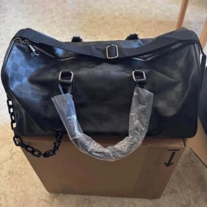 Lyxig svart weekendbag i rutigt mönster - Snygg svart weekendbag med diskret rutigt mönster och detaljer i silver. Väskan har både axelrem och handtag, samt en kedjedetalj i plast på sidan. Tillverkad i konstläder med dragkedja upptill och metallspänne. Perfekt för tex resor eller gymmet.  Mått: 43cm lång 21cm hög 18cm djup