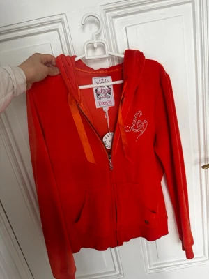 Röd hoodie med dragkedja från Lipsy - Snygg röd hoodie från Lipsy med dragkedja framtill och huva. Hoodien har orangea band i huvan och ett glittrigt 'L' i strass på bröstet. Mjuk och skön, perfekt för en avslappnad stil. Praktiska fickor framtill och ribbade muddar.