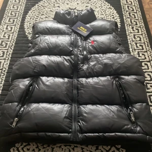 Svart dunväst från Polo Ralph Lauren - Snygg svart dunväst från Polo Ralph Lauren med klassisk röd logga broderad på bröstet. Västen har hög krage, två dragkedjefickor och quiltad design. Perfekt för lager-på-lager och streetstyle. Materialet är glansigt och ger en modern look.