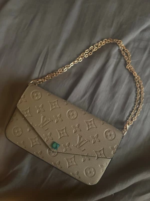 Beige kuvertväska - Snygg beige kuvertväska med klassiskt monogrammönster i präglat läder. Väskan har guldfärgad kedjerem och tryckknapp med turkos detalj. Invändigt finns ett fack med dragkedja för småsaker.