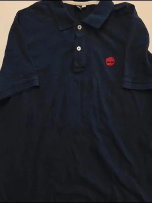 Mörkblå pikétröja från Timberland - Säljer en klassisk mörkblå pikétröja från Timberland med röd broderad logga på bröstet. Tröjan har en super bra fit med korta ärmar, krage och knäppning med två knappar och jättefin färg. Perfekt för en stilren och avslappnad look. STORLEK M  Bröstmått: ca 52 cm Längdmått: ca 68 cm