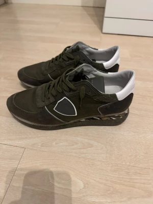 Philippe Model gröna sneakers - Snygga sneakers från Philippe Model i storlek 40 i mörkgrönt tyg och mocka med vita detaljer vid hälen. Skorna har en klassisk låg modell, svarta och vita detaljer på sulan samt märkets logga på sidan. Perfekta för dig som gillar streetstyle och vill sticka ut. Lådan till skorna finns inte och inte kvitto häller.