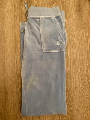  Juicy Couture  - Mysiga ljusblå mjukisbyxor från Juicy Couture i mjuk velour. Byxorna har bred resår i midjan, snörning och fickor fram samt bakfickor med guldknappar. Perfekta för chill dagar och har den klassiska Juicy-loggan broderad på ena fickan.