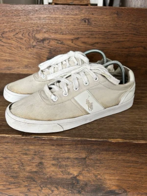 Polo Ralph Lauren Sneakers *42* - Snygga vita sneakers från Polo Ralph Lauren med klassisk låg profil och canvasmaterial. Skorna har vita snören, vit sula och den ikoniska Polo-loggan på sidan. Perfekta för en avslappnad och stilren look.
