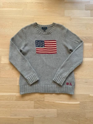 Grå stickad tröja Polo Ralph Lauren - Grå stickad tröja från Polo Ralph Lauren med amerikansk flagga i rött, vitt och blått på bröstet. Rund halsringning och ribbstickade muddar. RL-broderi i rött på nederkanten. Perfekt för dig som gillar klassisk och cool stil.