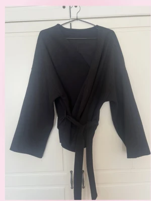 Svart omlottkappa med knytband - 💓En svart omlottkappa med breda ärmar och långt knytband i midjan. Kappan har en enkel och clean design utan synliga detaljer eller fickor. Materialet ser ut att vara mjukt och följsamt, perfekt för en avslappnad och trendig look.💕storlek M