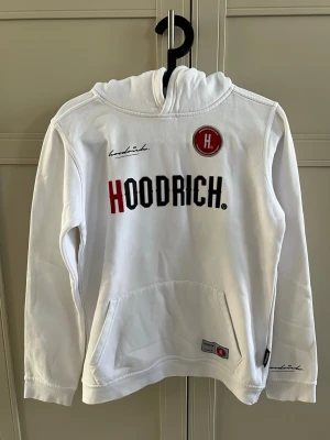 Vit hoodie från Hoodrich  - Snygg vit hoodie från Hoodrich med stor logga i svart och rött framtill och detaljerat tryck på ryggen. Hoodien har huva, känguruficka och ribbade muddar. Perfekt för dig som gillar streetwear och vill sticka ut med coola detaljer.
