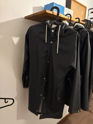 Svart regnjacka med huva - Snygg svart regnjacka med huva och vita dragsnören. Jackan har tryckknappar framtill, två fickor och långa ärmar. Tillverkad i vattenavvisande material, perfekt för regniga dagar. Enkel och stilren design som passar till det mesta.