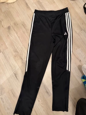 Svarta Adidas träningsbyxor - Säljer ett par svarta träningsbyxor från Adidas med de klassiska tre vita ränderna längs benen. Byxorna har resår i midjan och är tillverkade i ett lätt och stretchigt material som passar perfekt för sport och träning.