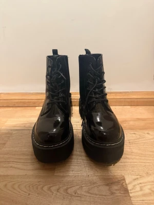 Svarta lackade boots - Säljer ett par svarta stövletter i glansigt lackmaterial med kraftig platåsula och snörning framtill. De har en chunky siluett och vita sömmar längs sulan för en cool kontrast. Perfekta för dig som vill ha en edgy och trendig look.