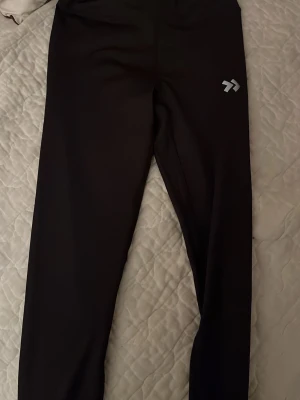 Svarta leggings med logga - Säljer ett par svarta leggings med hög midja och en liten vit logga på vänster lår. De är stretchiga och sitter tight, perfekta för träning eller chill. Materialet känns mjukt och följsamt.