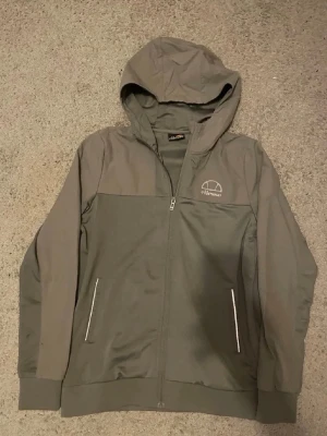 Grå hoodie från Ellesse med dragkedja - Snygg grå hoodie från Ellesse med dragkedja framtill och två fickor med vita detaljer. Hoodien har en klassisk huva och Ellesse-logga på bröstet. Tillverkad i ett mjukt och bekvämt material, perfekt för en avslappnad stil. Jag är 170cm lång och den sitter lite tajt men ändå bra och snyggt 