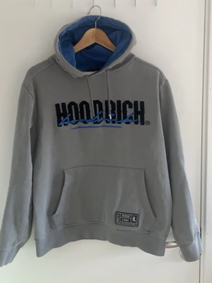 Grå Hoodrich hoodie med blå huva - Snygg grå hoodie från Hoodrich med blå insida på huvan och stor logga broderad i svart och blått framtill. Klassisk känguruficka och ribbade muddar. Perfekt för en avslappnad streetwear-look.