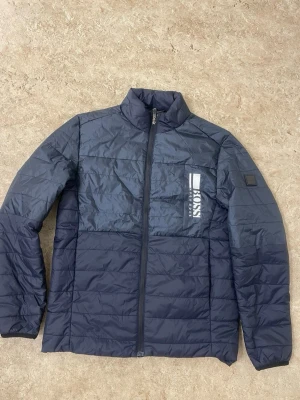 Mörkblå dunjacka från Boss - Snygg mörkblå dunjacka från Boss med hög krage och dragkedja framtill. Jackan har ett stort Boss-logotyptryck på bröstet och quiltad design som ger en modern look. Perfekt för kalla vinterdagar och enkel att matcha med olika outfits.