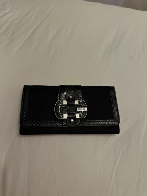 Svart plånbok från Guess med spänne - Snygg svart plånbok från Guess med ett stort metallspänne framtill och präglade detaljer. Plånboken har en stilren rektangulär form, mönstrad yta med Guess-loggor och en dragkedja på baksidan. 