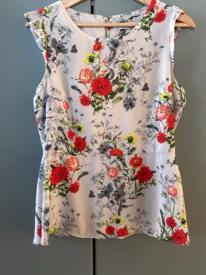 Blommig ärmlös topp med volang - Snygg ärmlös topp med volangkant och blommigt mönster i rött, grönt och gult på vit botten. Toppen har rund halsringning och dragkedja i ryggen. Perfekt för dig som vill ha en färgglad och somrig look.