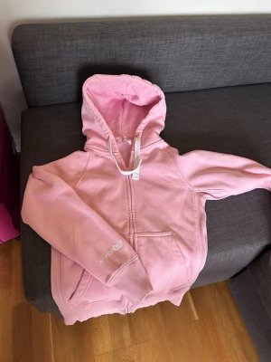 Ljusrosa hoodie med dragkedja - Mysig ljusrosa hoodie Russemerch med dragkedja framtill och två fickor. Har en stor huva med vita snören och broderad logga på ärmen. Tillverkad i mjukt bomullsmaterial med fleece på insidan för extra komfort. Finns en liten fläck mittpå bredvid dragkedjan. Syns på närbild. 