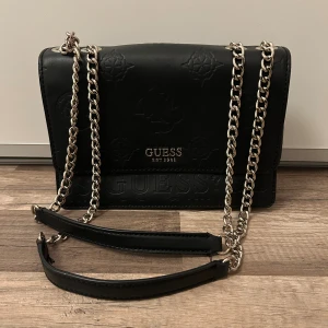 Svart axelremsväska från Guess  - Svart axelremsväska med präglad logga och silverfärgad kedja som axelrem från Guess. Väskan har flera fack, magnetlås och en dragkedja på baksidan. Observera att det förekommer ytterst små fläckar på väskan som nog går att ta bort och är inget som påverkar väskans funktion, se bilderna. Annars är väskan i mycket bra skick. 