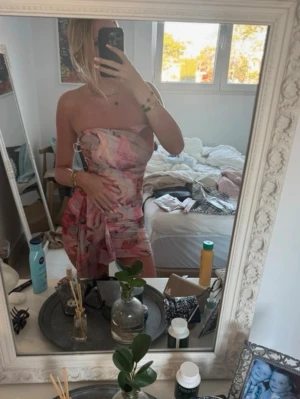 Rosa mönstrad axelbandslös klänning - Trendig kort axelbandslös klänning i rosa och ljusa toner med abstrakt mönster. Klänningen har en omlott-detalj på sidan och är figurnära med en snygg drapering. Perfekt för sommarens alla tillfällen.