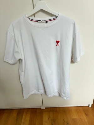 Vit t-shirt från Ami Paris med hjärtdetalj - Kostar 1200kr nytt- Vit t-shirt från Ami Paris med röd hjärtdetalj och bokstaven A broderad på bröstet. Klassisk rund halsringning och korta ärmar. Tillverkad i mjuk bomull för en skön känsla. Diskret AM-logga i nacken. 