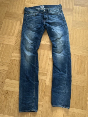 Blå raka jeans från Edwin - Säljer ett par klassiska blå jeans från Edwin med raka ben och snygg slitning på låren och knäna. Jeansen har fem fickor, gylf med dragkedja och är tillverkade i slitstark denim. Perfekta för en avslappnad och trendig look.