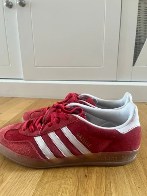 Adidas Gazelle röda sneakers - Säljer ett par klassiska Adidas Gazelle sneakers i rött mocka med vita detaljer och de ikoniska tre ränderna på sidan. Skorna har lågt skaft, vita snören och gummisula. Perfekta för dig som vill ha en retro och sportig look.