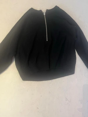 Svart ribbad tröja med dragkedja - Svart ribbad tröja med lång ärm och silverfärgad dragkedja framtill. Tröjan har en rund halsringning och bred mudd nedtill. Perfekt för en stilren och enkel look. Andvänt ganska mycket men bra skick lite nopprig men är lätt att ta bort 