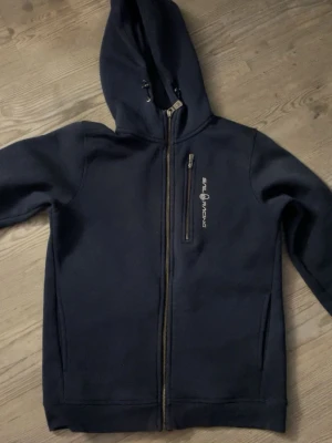 Marinblå hoodie från Sail Racing - Snygg marinblå hoodie från Sail Racing med dragkedja framtill och en extra bröstficka. Tillverkad i mjukt och bekvämt material, perfekt för chill dagar. 💓