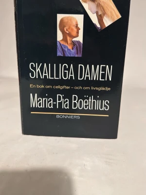 Skalliga Damen av Maria-Pia Boëthius - En svart inbunden bok med titeln 'Skalliga Damen' av Maria-Pia Boëthius. Omslaget har två bilder och text i vitt och gult. Boken handlar om cellgifter och livsglädje och ges ut av Bonniers.
