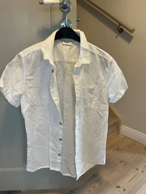 Vit kortärmad linneskjorta från Kappahl - Säljer en vit skjorta från Kappahl i storlek 164. Skjortan har korta ärmar, klassisk krage och knappar framtill. Materialet är linne och den har en bröstficka..
