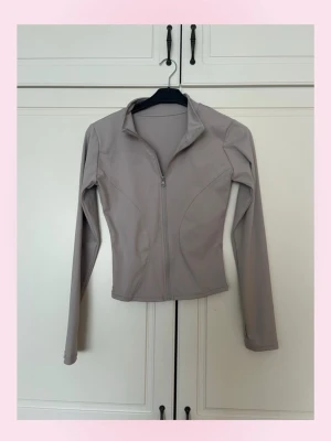 Beige grå sportjacka med dragkedja - 💗Snygg beige grå sportjacka med figurnära passform och hög krage. Jackan har långa ärmar och hel dragkedja framtill. Tillverkad i ett stretchigt och mjukt material som passar perfekt till träning eller som ett extra lager. Enkel och stilren design 💓