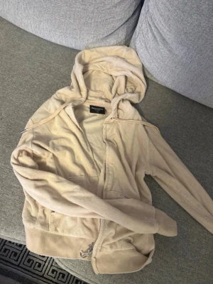 Beige velourhoodie från Juicy Couture - Mysig beige hoodie i velour från Juicy Couture med dragkedja framtill, huva med dragsko och broderad JC-logga på bröstet. Perfekt för en avslappnad och trendig look. Klassisk modell med ribbade muddar och fickor framtill. Betalning via vanlig swish 