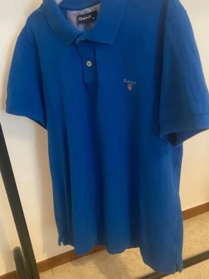 Blå pikétröja från GANT - Klassisk blå pikétröja från GANT med korta ärmar och krage. Tröjan har knappslå framtill och en liten broderad logga på bröstet. Perfekt till en avslappnad och stilren look.