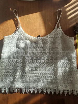 Vit spetsig linnetopp med tunna band - Fin vit topp med tunna axelband och spets. Den har ett fint mönster och en dekorativ kant längst ner. Använd få gånger. Perfekt till sommaren och lätt att matcha med jeans eller kjol.