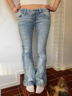 Bootcut ljusblå jeans med slitningar - Säljer ett par ljusblå bootcut jeans med slitningar på låret och knät. Jeansen har dekorativa bakfickor med knappar och mönstrade detaljer. Klassisk femficksmodell och låg midja, perfekta till en avslappnad stil. Skriv ifall frågor 