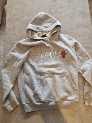 Grå Chicago Bulls hoodie från Jordan - Snygg grå hoodie från Jordan med Chicago Bulls-logga och texten 'Chicago' broderat på bröstet. Hoodien har stor känguruficka framtill, ribbade muddar och en mjuk insida. Perfekt för dig som gillar streetwear och basketstil.