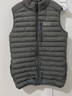 Grå dunväst från McKenzie - Snygg grå dunväst från McKenzie med quiltad design och hög krage. Västen har hel dragkedja framtill, en bröstficka med dragkedja och två sidofickor. Perfekt för lager på lager och håller dig varm under kyliga dagar.