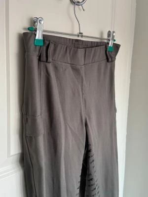 Grå/beige ridtights med ficka - Säljer ett par grå ridtights med hög midja och praktisk ficka på sidan. Tightsen har silikonmönster på insidan av benen för extra grepp i sadeln. Perfekta för ridning och träning, tillverkade i stretchigt funktionsmaterial.