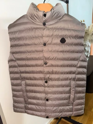 Grå dunväst från Moncler - Snygg grå dunväst från Moncler med hög krage och svarta knappar framtill. Västen har två fickor och Monclers logotyp på bröstet. Tillverkad i lätt, quiltad dun med en stilren och modern look.