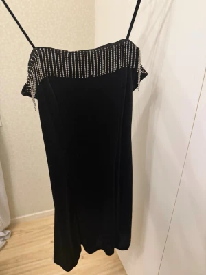 Svart sammetsklänning med strass - Supersnygg svart sammetsklänning med tunna axelband och glittriga strassdetaljer längs överdelen. Klänningen har en rak passform och är perfekt för dig som vill sticka ut. Materialet känns lyxigt och mjukt mot huden. Använd endast en gång på nyårsafton 