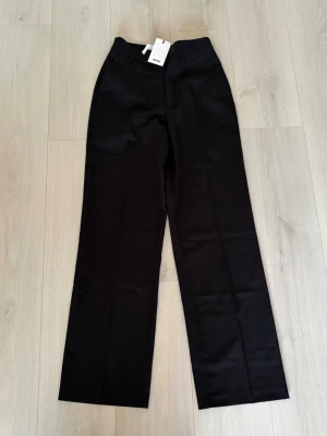 Svarta kostymbyxor från BikBok - Stilrena svarta kostymbyxor från BikBok med raka ben och klassisk passform. Byxorna har hällor för skärp och stängs med knapp och dragkedja. Perfekta för en clean och snygg look. Materialet är mjukt och har en lätt glansig finish.