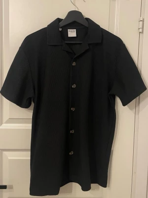 Svart ribbad skjorta från Selected Homme - Stilren svart skjorta från Selected Homme med korta ärmar och ribbad struktur. Skjortan har klassisk krage och knappar framtill, perfekt för en clean och modern look. Tillverkad i mjuk bomull för skön känsla.